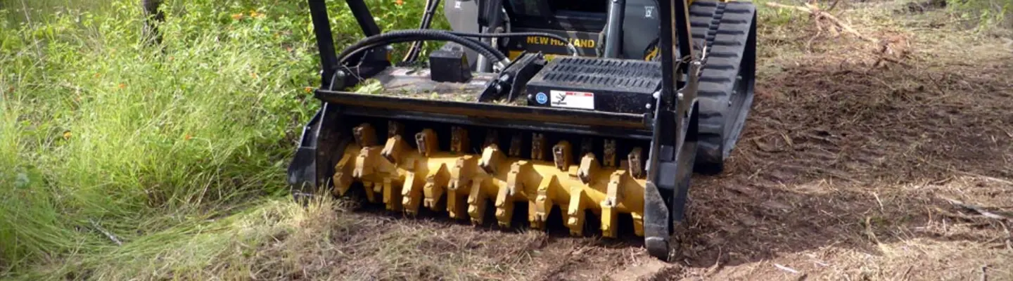 Large machine trimming overgrown grass in an open field near highway barriers to support roadside safety and visibility.