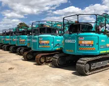 A row of construction equipment lined up on a worksite, inspected and ready for use, with safety barriers in the background.
